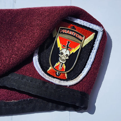 Maroon fabric with a black strap and a decorative badge featuring a skull and crossbones on a white background