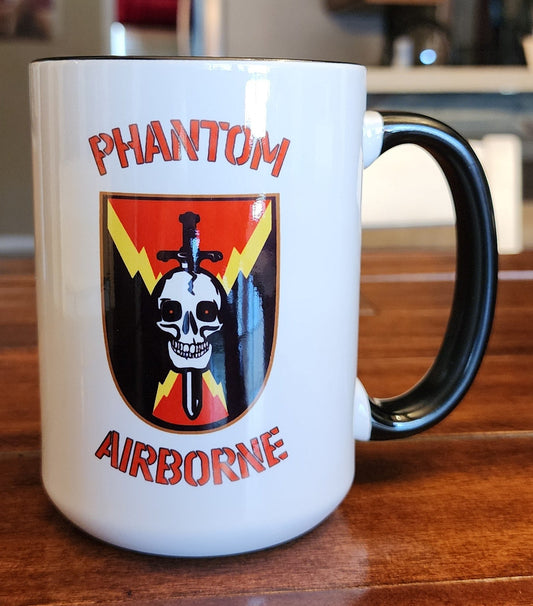 White mug with 'Phantom Airborne' design on a wooden table in a kitchen.